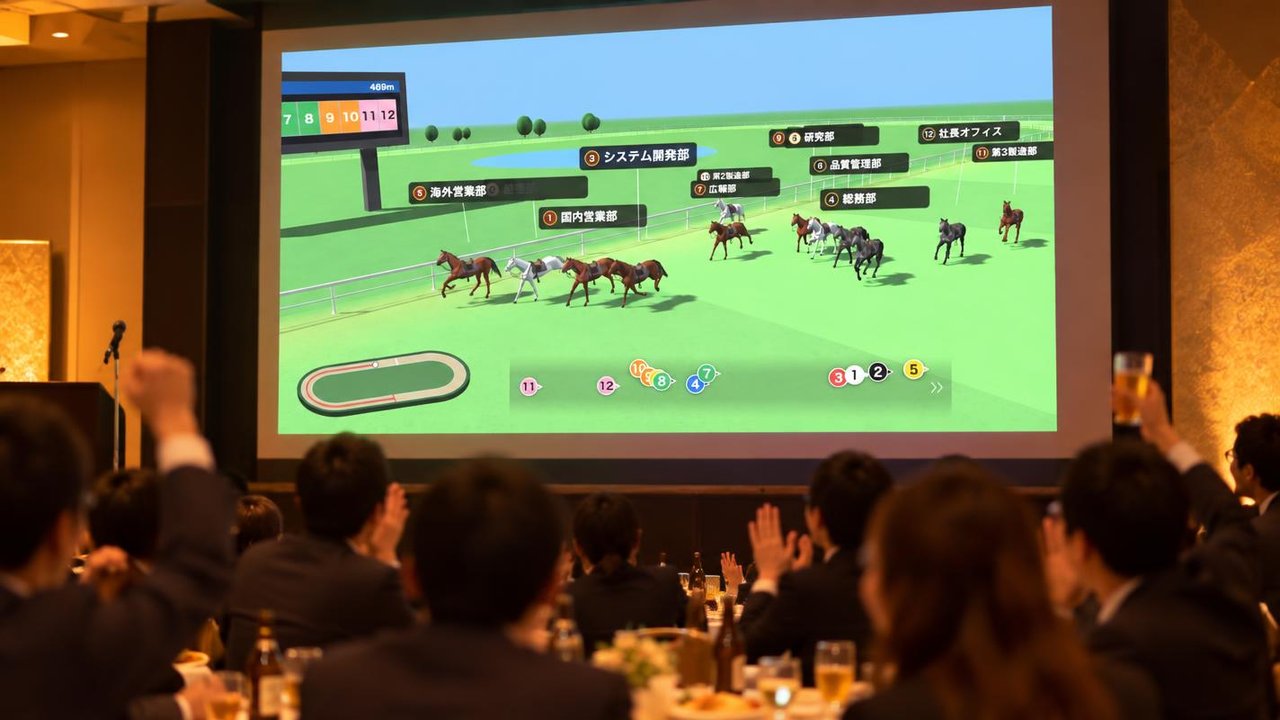 忘年会の会場で、スクリーンに映した格付けダービーの3Dレースを参加者が盛り上がりながら見ている様子。出走馬の名前は部署名になっている